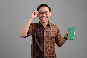 Cheerful young Asian man wearing batik shirt showing blank screen smartphone and touching glasses isolated on grey background