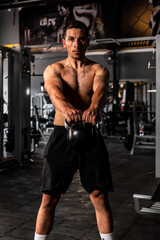 Fototapeta na wymiar A very muscular Latino man standing up straight lifting a kettlebell with great ease. Latino man without a shirt training in the gym. High quality photo