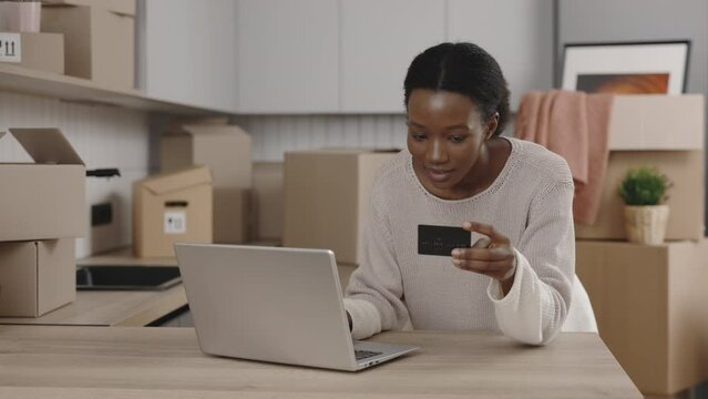 African american woman pays for purchases from a new apartment via the Internet using a card and a laptop at new home. Female is paying with credit card. Relocation. People and technology concept.