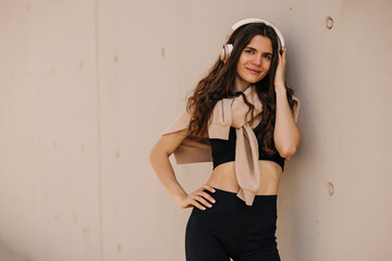View of european attractive woman looking to camera in headphones on wall background. Wearing black top, leggings, tied hoodie at shoulders. Street style, technology concept