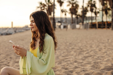 The side view of european young woman chatting and looking with smile to phone. Wearing green costume sitting at beach. Use telephone technology concept 