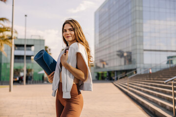 Image of pretty caucasian young woman staying on city background holding blue mat. Looking and smiling to camera, in brown sportswear. Lifestyle, street style concept 