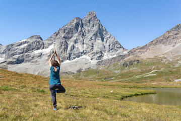 Naklejka premium Rear view woman practising yoga on a green meadow at a mountain lake shore with Matterhorn in front of her in Italian Alps. Yoga in nature.