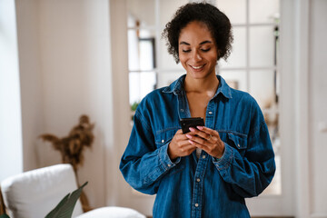 Young woman wearing denim shirt smiling and using cellphone at home