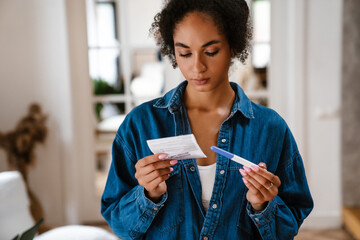Young black woman holding pregnancy test and instruction at home