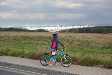 A little girl on her bicycle looks at the landscape. A child on a bicycle is stopped and looks at the landscape in front of her. In the distance is a large city.