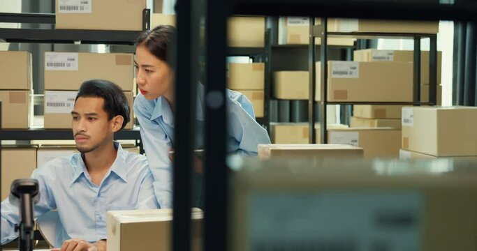 Happy Asian man and lady partner wear formal shirt sit in front of desk computer and barcode machine scanner discuss stock detail customer online data delivery at warehouse. Startup business concept.