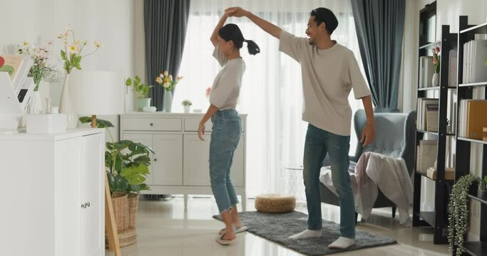 Happy young Asian couple enjoy cheerful time dancing together, celebrating relocation or anniversary in living room at home on holiday. Smile young husband and wife moving for favorite song.