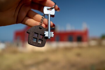 A woman's hand holds a house key against the backdrop of a house under construction. Real estate agent. Buying a house, apartment.
