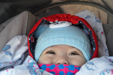 The child is sitting in a stroller with a pink scarf and a blue hat, only the eyes and nose are visible.