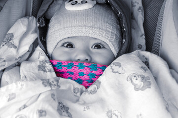 The child is sitting in a stroller with a pink scarf and a blue hat, only the eyes and nose are visible.