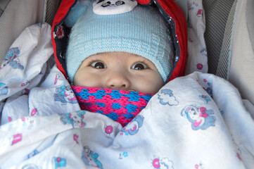 The child is sitting in a stroller with a pink scarf and a blue hat, only the eyes and nose are visible.