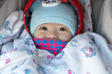 The child is sitting in a stroller with a pink scarf and a blue hat, only the eyes and nose are visible.