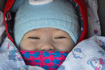 The child is sitting in a stroller with a pink scarf and a blue hat, only the eyes and nose are visible.