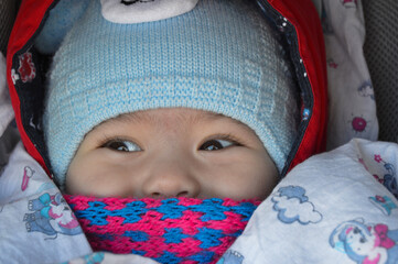 The child is sitting in a stroller with a pink scarf and a blue hat, only the eyes and nose are visible.
