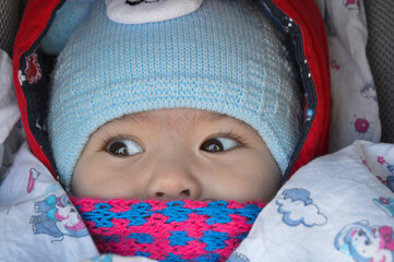 The child is sitting in a stroller with a pink scarf and a blue hat, only the eyes and nose are visible.