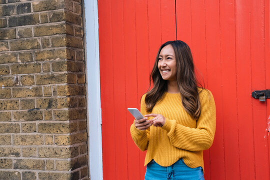 Happy Young Woman With Mobile Phone Standing In Front Of Red Door