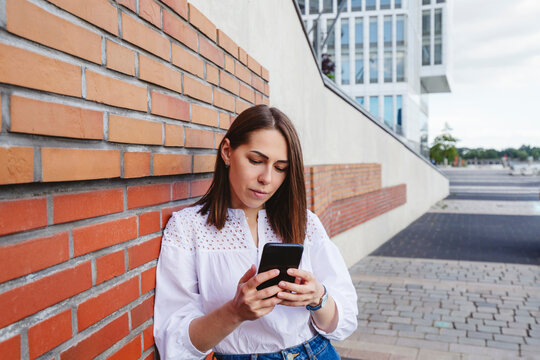 Serious woman looking at mobile phone leaning on brick wall