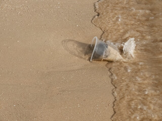 Close up top view of a Waste transparent plastic cup on the sand beach, a moment before rushed by sea wave, with copy space.