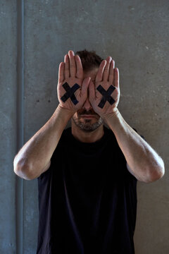 Man covering eyes with hands having cross marking in front of wall