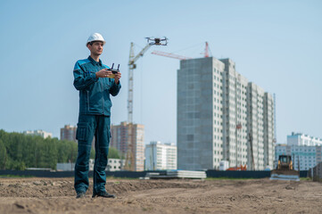 A man in a helmet and overalls controls a drone at a construction site. The builder carries out technical oversight.
