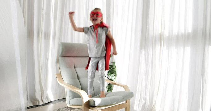Little girl in a superhero costume runs into an armchair and jumps, rejoicing, raising her hand in a bright sunny room