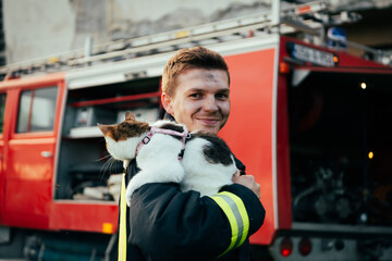 Close-up portrait of heroic fireman in protective suit and red helmet holds saved cat in his arms....