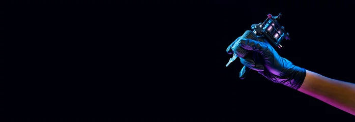 Foto op Aluminium Closeup of tattooer master's hand in black glove holding machine for making tattoo art on body isolated on dark background in neon light. © master1305