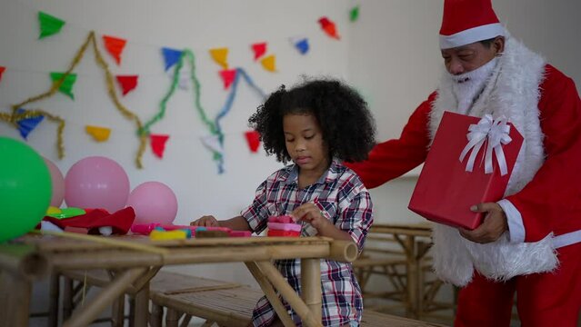 Little Kid sitting on the bamboo table concentrate play plasticine Santa come from behind and give gift box
