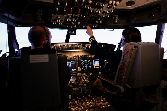 Woman Copilot Assisting Captain To Takeoff And Fly Airplane, Using Buttons On Dashboard Command In Pilot Cockpit. Airliners Flying Plane Jet With Navigation Windscreen And Control Panel.