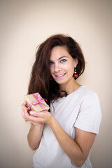 pretty young woman with beautiful long hair is holding a small christmas gift in her hand and has earrings that look like christmas tree baubles standing in front of light brown background