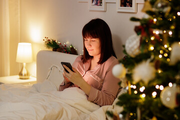 young woman using smartphone in decorated bedroom with christmas tree