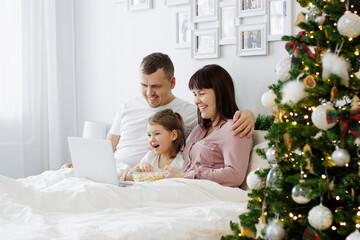 christmas, family and love concept - happy family sitting on the bed and watching movie near christmas tree