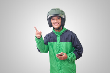 Portrait of Asian online taxi driver wearing green jacket and helmet holding mobile phone and pointing to empty space with finger. Isolated image on white background