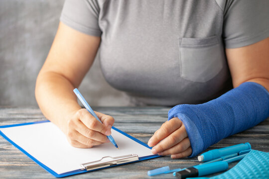Woman with  blue cast on her arm writes insurance with a pen on paper on the  grey background. Girl has a broken hand wearing