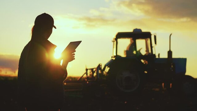 Silhouette woman uses specialized app on a digital tablet PC on background of working tractor in field. Remote control machinery equipment on farmland. Concept optimization of work of small business.
