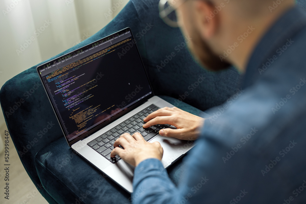 Poster young man in denim jacket coding on laptop while sitting on blue couch, remote networking concept