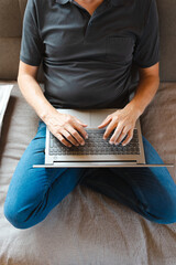Ein Mann über 50 sitzt im Schneidersitz auf einer Couch und tippt auf einem Laptop. Zuhause.