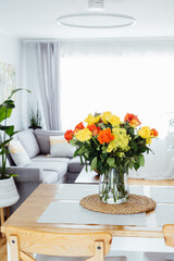Kitchen counter table with focus on vase with yellow and orange roses on it with blurred background of modern cozy living room with couch and green plants. Open space home interior design. Copy space