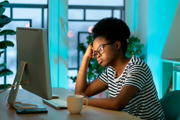 Pensive African woman wearing glasses works freelancer sitting at table at home. Kinky-haired entrepreneur thinks about new business project looking at monitor of computer against potplants by window