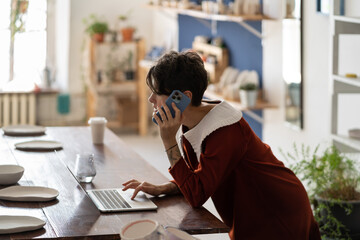 Young woman pottery business owner using laptop and talking with client by phone while working in workshop. Female artisan making money selling ceramics. Ceramic shop manager checking online order