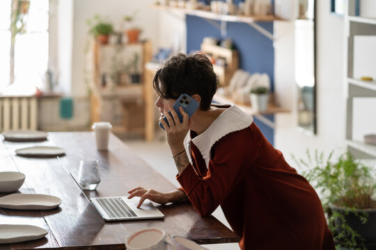 Young woman pottery business owner using laptop and talking with client by phone while working in workshop. Female artisan making money selling ceramics. Ceramic shop manager checking online order
