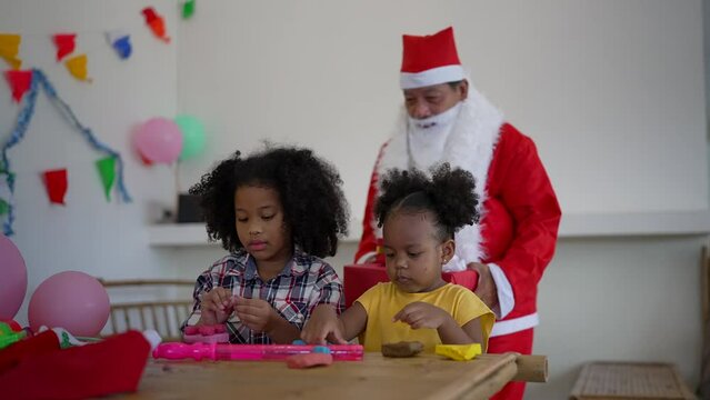 2 Kid sister and daughter sitting on the bamboo table concentrate play plasticine Santa come from behind and give gift box to them
