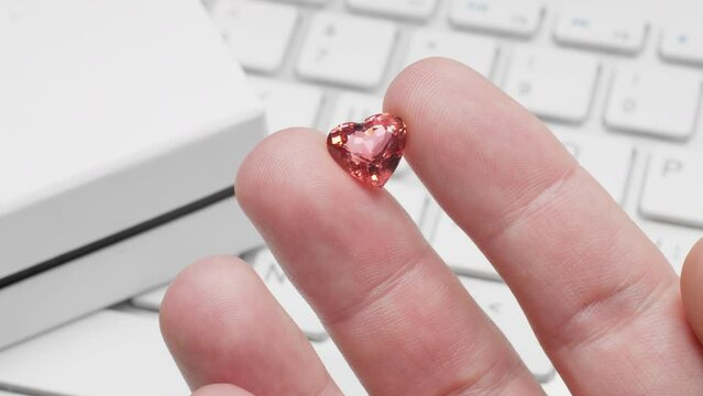 Heart shaped tourmaline gem in hand of gemologist or jeweller. Online jewelry shopping for birthday, Valentine Day or other holiday. Male hand holding red tourmaline, macro shot with box and keyboard.