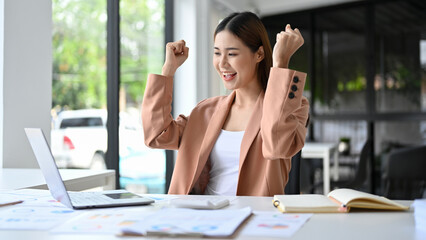 Successful Asian businesswoman celebrating at her office desk. achieve goal.