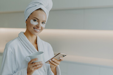 Horizontal shot of glad beautiful womanwith healthy skin cares about undereye skin applies moisturising patches poses in bathrobe uses mobile phone for surfing social networks drinks coffee.