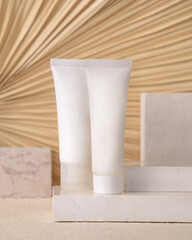 White plastic cream tubes on travertine stone against dry beige palm leaf close up, cosmetic mockup