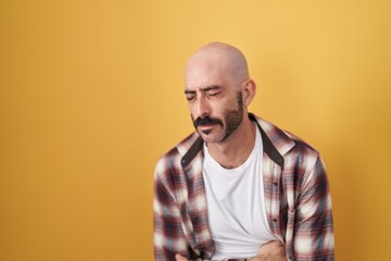 Hispanic man with beard standing over yellow background with hand on stomach because nausea,...