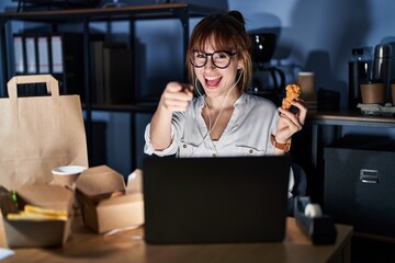 Young beautiful woman working using computer laptop and eating delivery food pointing fingers to camera with happy and funny face. good energy and vibes.