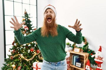 Redhead man with long beard wearing christmas hat by christmas tree celebrating mad and crazy for success with arms raised and closed eyes screaming excited. winner concept